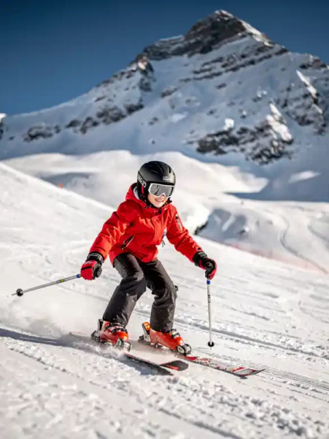 Beginnende skiër in rode jas daalt voorzichtig een brede piste af met besneeuwde Alpentoppen op de achtergrond.
