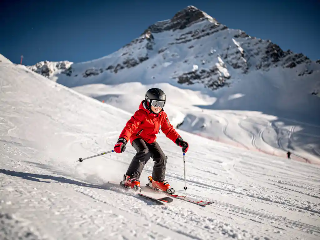 Beginnende skiër in rode jas daalt voorzichtig een brede piste af met besneeuwde Alpentoppen op de achtergrond.