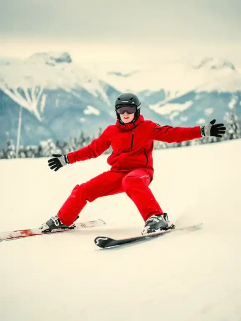 Beginnende skiër in rood jack met snowploegtechniek op verzorgde alpine piste, met besneeuwde bergtoppen op de achtergrond.