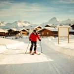 Beginner skiër in felrood jack daalt voorzichtig een zachte oefenpiste af, met skischoolbord en Alpendorp op de achtergrond.