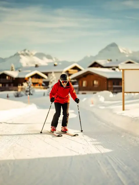 Beginner skiër in felrood jack daalt voorzichtig een zachte oefenpiste af, met skischoolbord en Alpendorp op de achtergrond.