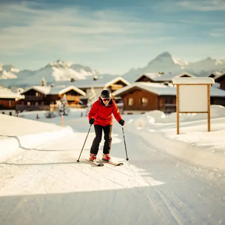 Beginner skiër in felrood jack daalt voorzichtig een zachte oefenpiste af, met skischoolbord en Alpendorp op de achtergrond.