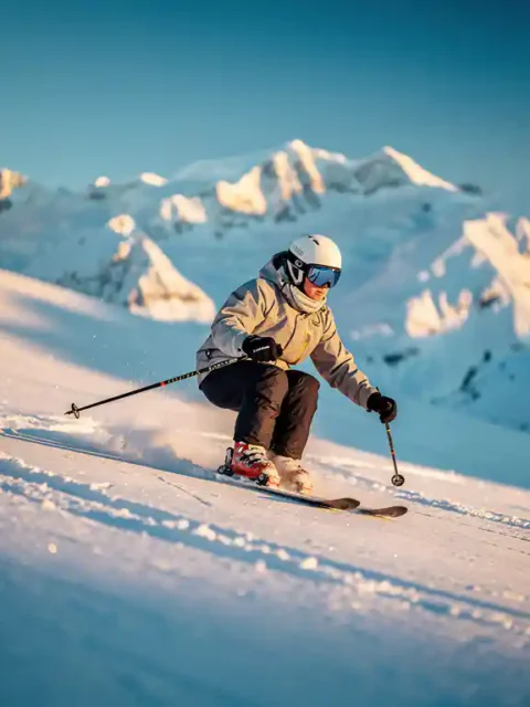 Beginnende skiër maakt eerste gesneden bocht op brede Alpijnse piste, met gouden ochtendlicht en kobaltblauwe lucht op de achtergrond.