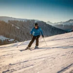 Beginnende skiër in felblauwe jas daalt voorzichtig een groene piste af, omgeven door besneeuwde Alpiene hellingen met dennenbomen.