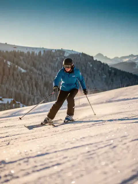 Beginnende skiër in felblauwe jas daalt voorzichtig een groene piste af, omgeven door besneeuwde Alpiene hellingen met dennenbomen.