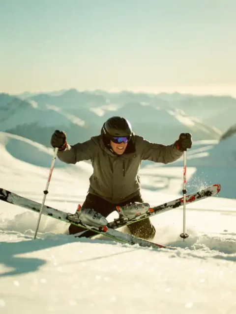 Beginnende skiër gevallen in poedersneeuw op zonnige alpiene helling, opstaan met skistokken in glinsterende witte bergen.