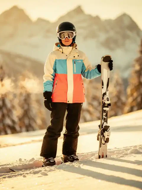 Beginnende skiër in felgekleurd huurjack op zachte besneeuwde helling, met korte brede ski's in verse poedersneeuw, Alpiene achtergrond.