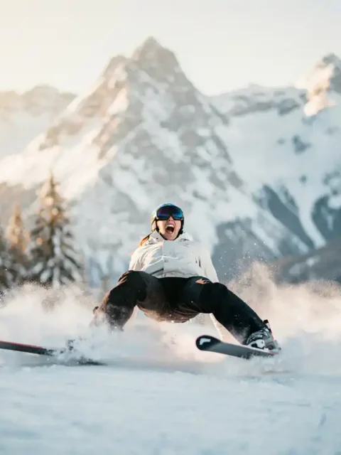 Beginnende skiër op besneeuwde Alpenhelling die achterover leunt met uitwaaierende ski's, mislukte pizzapunt-houding, dennenbomen op de achtergrond.