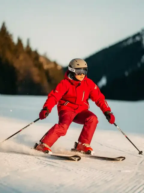 Beginnende skiër in rood jack voert een ploegschaar-bocht uit op een verzorgde blauwe piste, skitoppen in V-vorm in witte sneeuw.