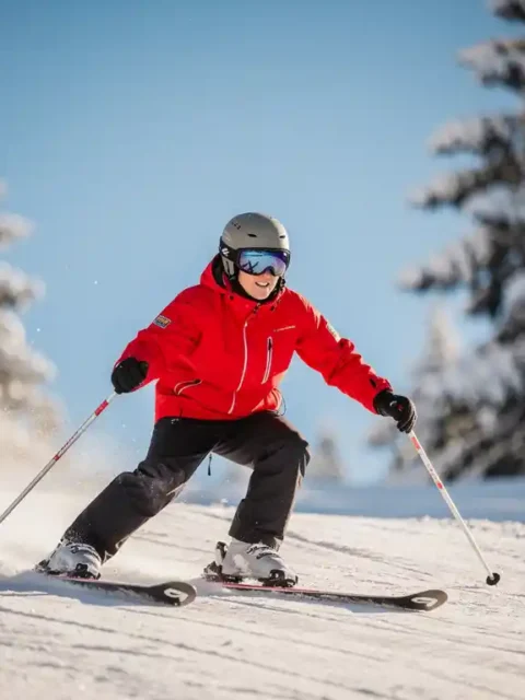 Beginner skiër in felrood jack maakt een ploegbocht op een zacht glooiende, verzorgde piste met besneeuwde dennenbomen op de achtergrond.