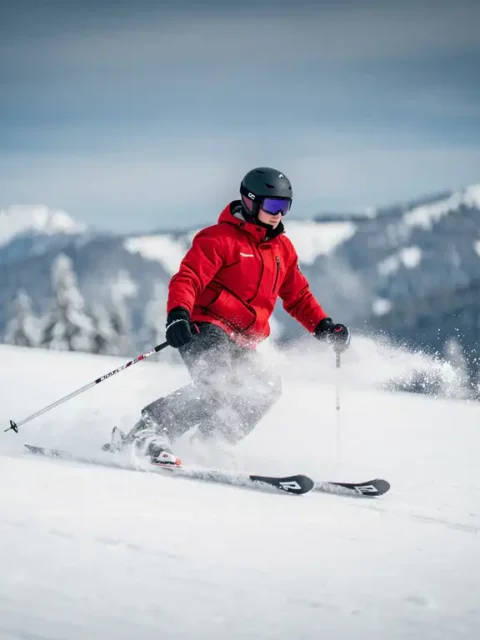 Beginner skiër op verzorgde alpine piste in rood jack, maakt onzekere bocht met skis naar buiten, verse sneeuw spat omhoog.