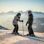 Nerveuze beginner op ski's met instructeur bovenaan een zachte piste, zonnige Alpiene bergpanorama op de achtergrond.