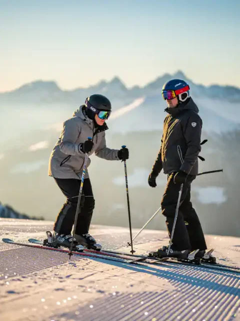 Nerveuze beginner op ski's met instructeur bovenaan een zachte piste, zonnige Alpiene bergpanorama op de achtergrond.