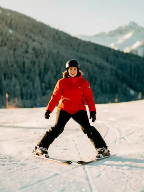 Beginnende skiër in rood jack in pizzawig-positie op verzorgde alpine piste, omringd door sparrenbergen en gouden winterzon.