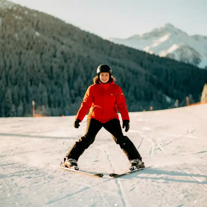 Beginnende skiër in rood jack in pizzawig-positie op verzorgde alpine piste, omringd door sparrenbergen en gouden winterzon.