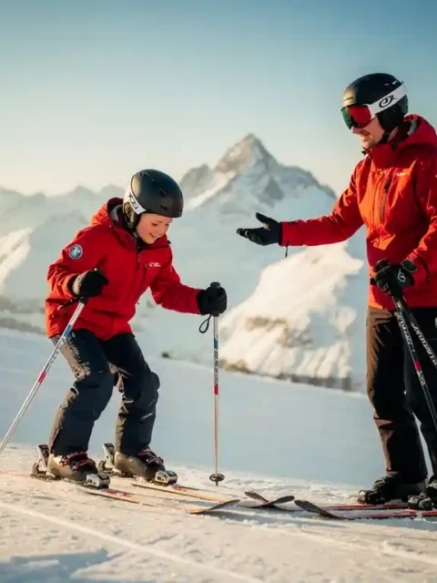 Beginnende skiër in rood jack op zachte sneeuwhelling, begeleid door skileraar met aanmoedigend gebaar, Alpen panorama op de achtergrond.
