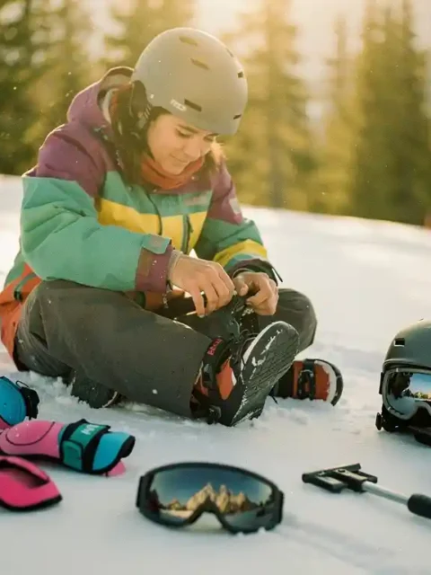 Beginner snowboarder adjusting bindings on snowy slope with helmet, goggles, wrist guards and safety gear scattered around