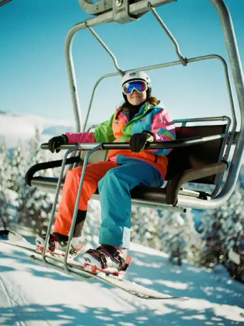 Beginner snowboarder in bright gear riding chairlift above snow-covered mountains and pine trees on sunny day