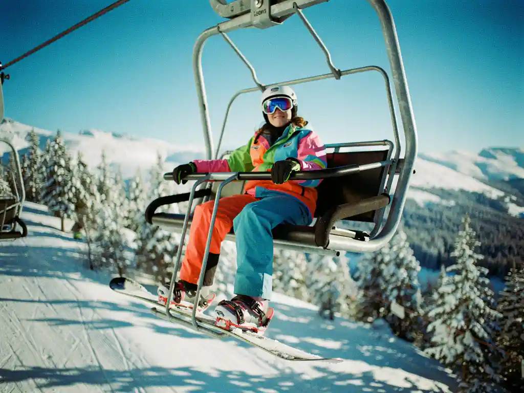 Beginner snowboarder in bright gear riding chairlift above snow-covered mountains and pine trees on sunny day