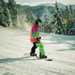 Beginner snowboarder in bright winter gear descending gentle green slope with trail markers and snow-covered pine trees.