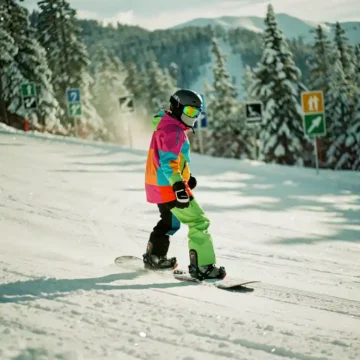 Beginner snowboarder in bright winter gear descending gentle green slope with trail markers and snow-covered pine trees.