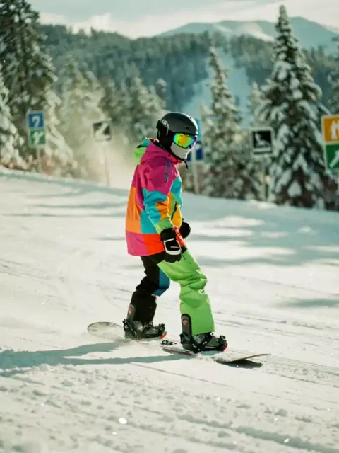 Beginner snowboarder in bright winter gear descending gentle green slope with trail markers and snow-covered pine trees.