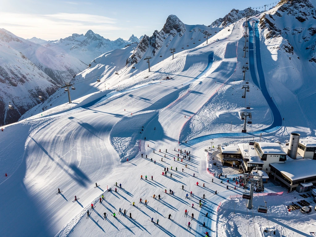 Aerial view of Isola 2000 ski resort with groomed slopes, ski lesson groups, and snow-covered Alpine peaks in morning sunlight.