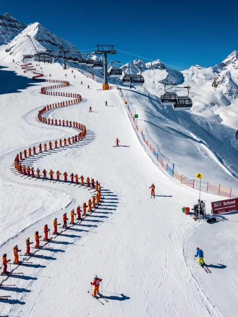 Aerial view of Kaltenbach ski slopes with instructors leading colorful groups down groomed Alpine runs under blue skies.
