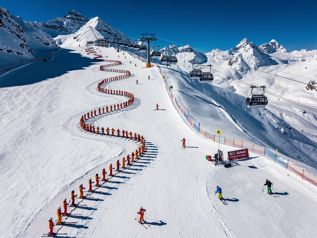 Aerial view of Kaltenbach ski slopes with instructors leading colorful groups down groomed Alpine runs under blue skies.