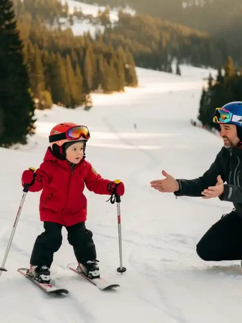 Jong kind in rode skijas houdt skipoles vast op een zachte beginnersskihelling, met een geduldige ski-instructeur ernaast.
