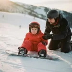 Kind van 6 jaar in rode winterkleding leert snowboarden van instructeur op besneeuwde bergpiste in zonlicht