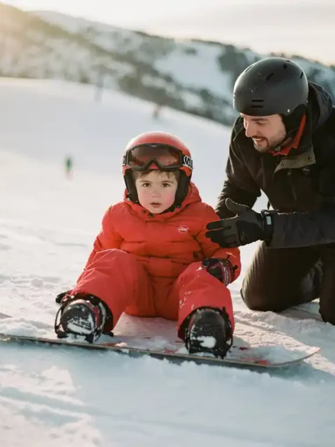 Kind van 6 jaar in rode winterkleding leert snowboarden van instructeur op besneeuwde bergpiste in zonlicht