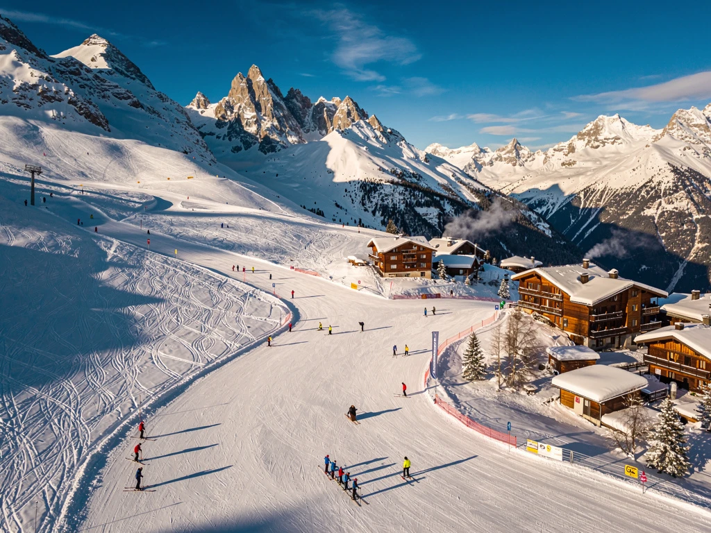 Aerial view of Les Contamines-Montjoie ski resort with groomed slopes, colorful skiers, and snow-capped French Alps peaks.