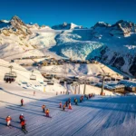 Aerial view of Les Deux Alpes ski resort with groomed slopes, colorful skiers, ski lifts, and snow-covered Alpine peaks.