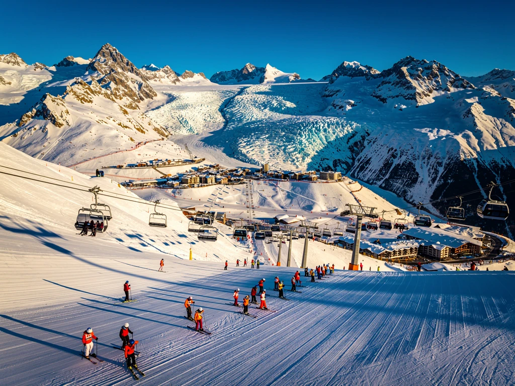 Aerial view of Les Deux Alpes ski resort with groomed slopes, colorful skiers, ski lifts, and snow-covered Alpine peaks.
