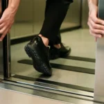 Persoon stapt voorzichtig lift in, één voet binnen en één buiten, handen aan deurkozijn voor stabiliteit