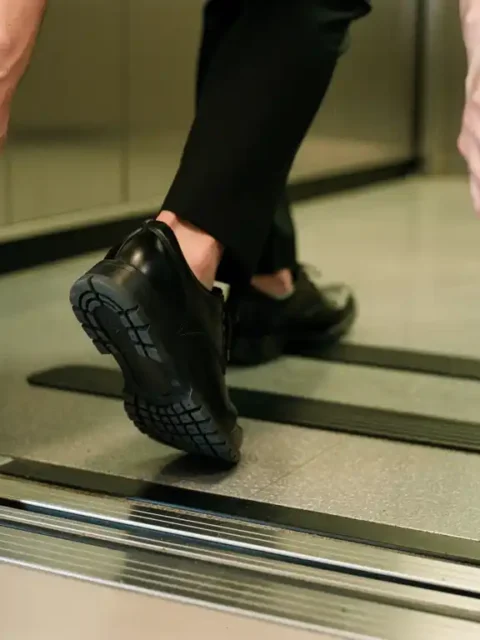 Persoon stapt voorzichtig lift in, één voet binnen en één buiten, handen aan deurkozijn voor stabiliteit