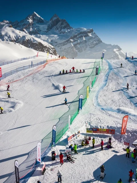 Aerial view of Orelle ski slopes with colorful skiers and instructors on groomed runs beneath snow-capped Alpine peaks.