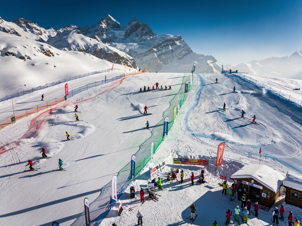 Aerial view of Orelle ski slopes with colorful skiers and instructors on groomed runs beneath snow-capped Alpine peaks.