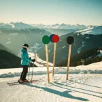 Skiër bij pistekruising met groene, blauwe, rode en zwarte pistenmarkeringen in verse sneeuw, met Alpien dal op de achtergrond.