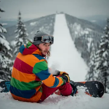 Snowboardeur nerveux en équipement d'hiver assis au bord d'une pente enneigée, regardant la montagne avec détermination