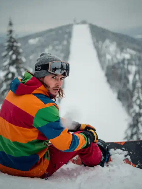 Snowboardeur nerveux en équipement d'hiver assis au bord d'une pente enneigée, regardant la montagne avec détermination