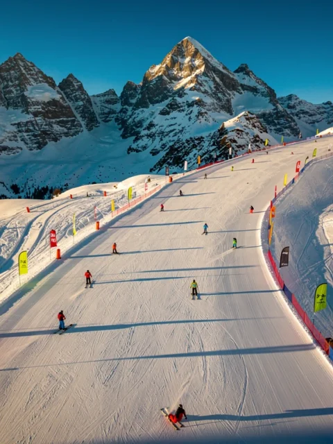 Aerial view of Schladming ski slopes with groomed powder snow trails, colorful skiers, and snow-capped Austrian Alpine peaks.