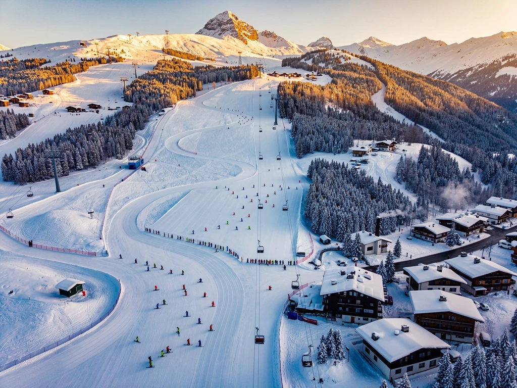 Aerial view of Schruns ski resort in Austrian Alps with ski lesson groups on groomed slopes during golden hour lighting.