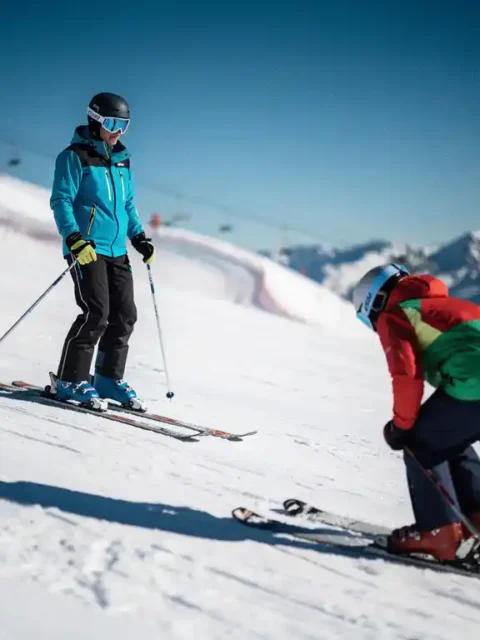Ski-instructeur met rechtopstaande houding op verzorgde alpinehelling, op de achtergrond een skiër met gebogen rug en ingezakte schouders.