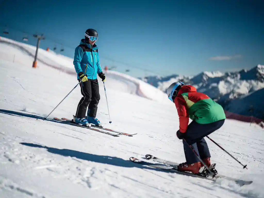 Ski-instructeur met rechtopstaande houding op verzorgde alpinehelling, op de achtergrond een skiër met gebogen rug en ingezakte schouders.