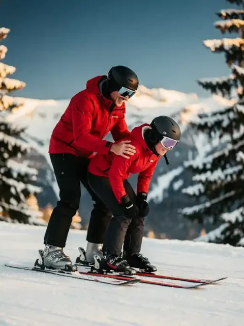 Ski-instructeur corrigeert kniebuiging en voorwaartse houding van leerling op besneeuwde piste, omgeven door dennenbomen.