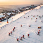 Aerial view of Super Besse ski resort slopes with skiers and red-jacketed instructors during golden hour sunset.