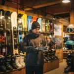 Freundlicher Mitarbeiter prüft Snowboard-Bindung in professionellem Ski-Shop mit organisierten Snowboards und Helmen