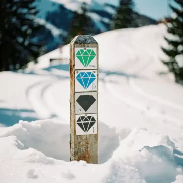 Wooden ski trail marker post with colored diamond difficulty symbols surrounded by fresh powder snow and pine trees